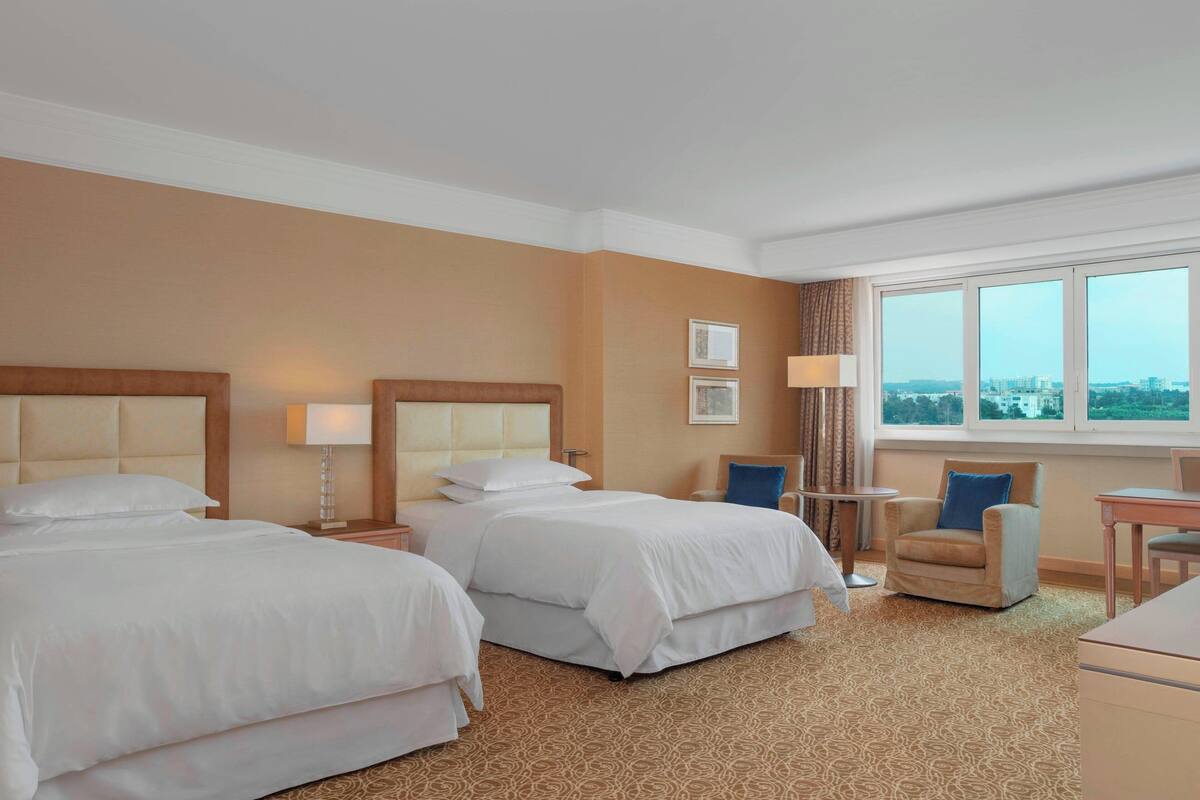 Ein Hotelzimmer mit zwei Betten, einem kleinen Tisch, einem Stuhl und einem Fenster mit Blick auf die Stadt.