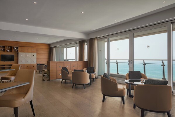 A modern interior with a wooden panel wall, a bar area, and seating arrangements facing large windows with a view of the sea.