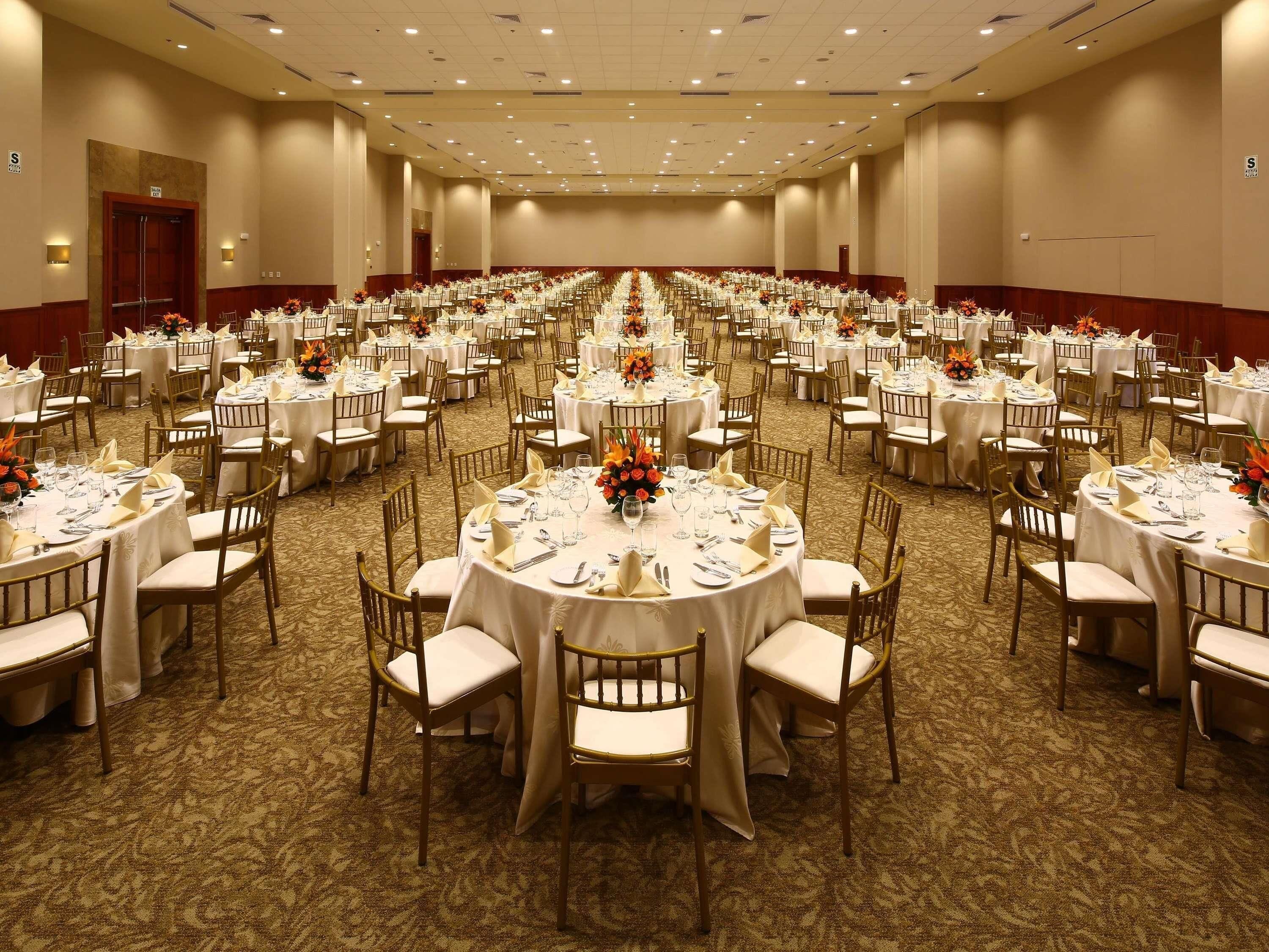 Un amplio salón de banquetes con mesas redondas, cada una con mantel blanco, sillas y centros de mesa florales.