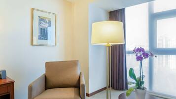 A hotel room with a chair, a floor lamp, a small table with a potted plant, a framed picture on the wall, and a large window with curtains.