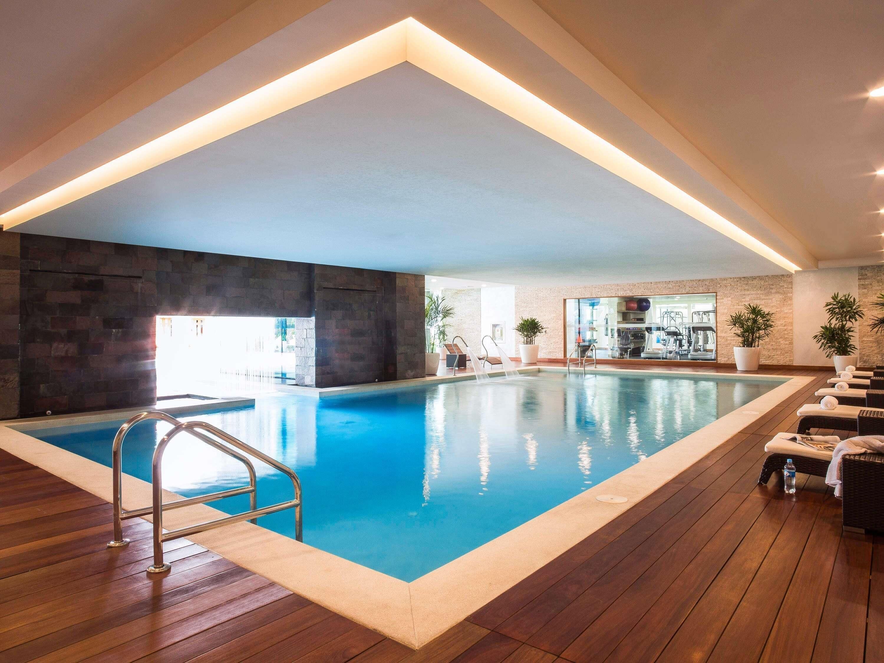 An indoor swimming pool with a wooden deck, metal railings, and potted plants.