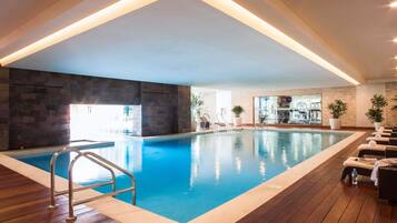 An indoor swimming pool with a wooden deck, metal railings, and potted plants.