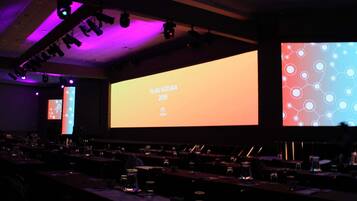 Un salón de conferencias con una pantalla grande mostrando 'PLAN NATURA 2019', varias mesas con sillas e iluminación en el techo.