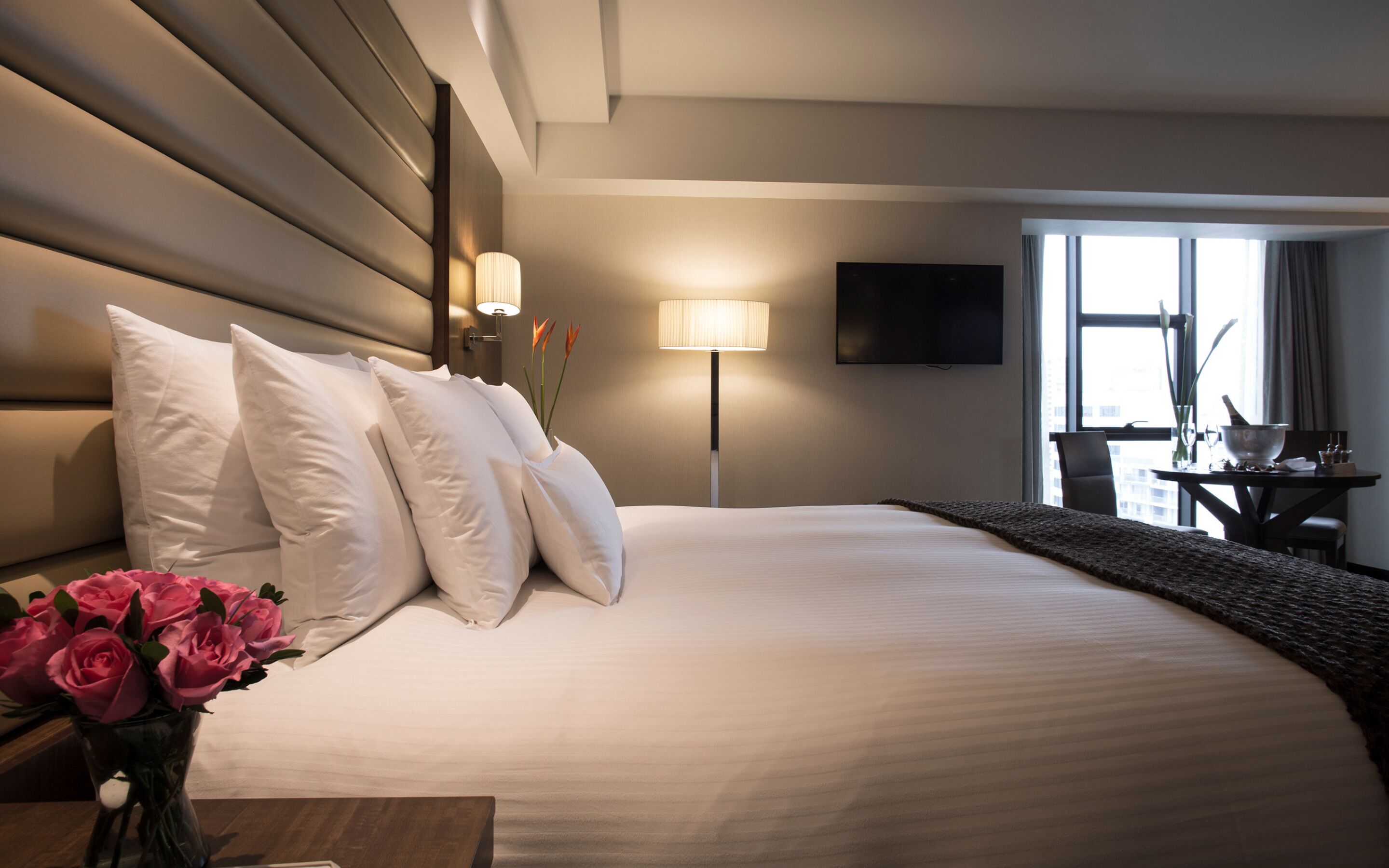 A hotel room with a large bed, white pillows, a bedside table with a vase of flowers, a lamp, and a TV on the wall.
