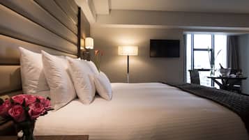 A hotel room with a large bed, white pillows, a bedside table with a vase of flowers, a lamp, and a TV on the wall.