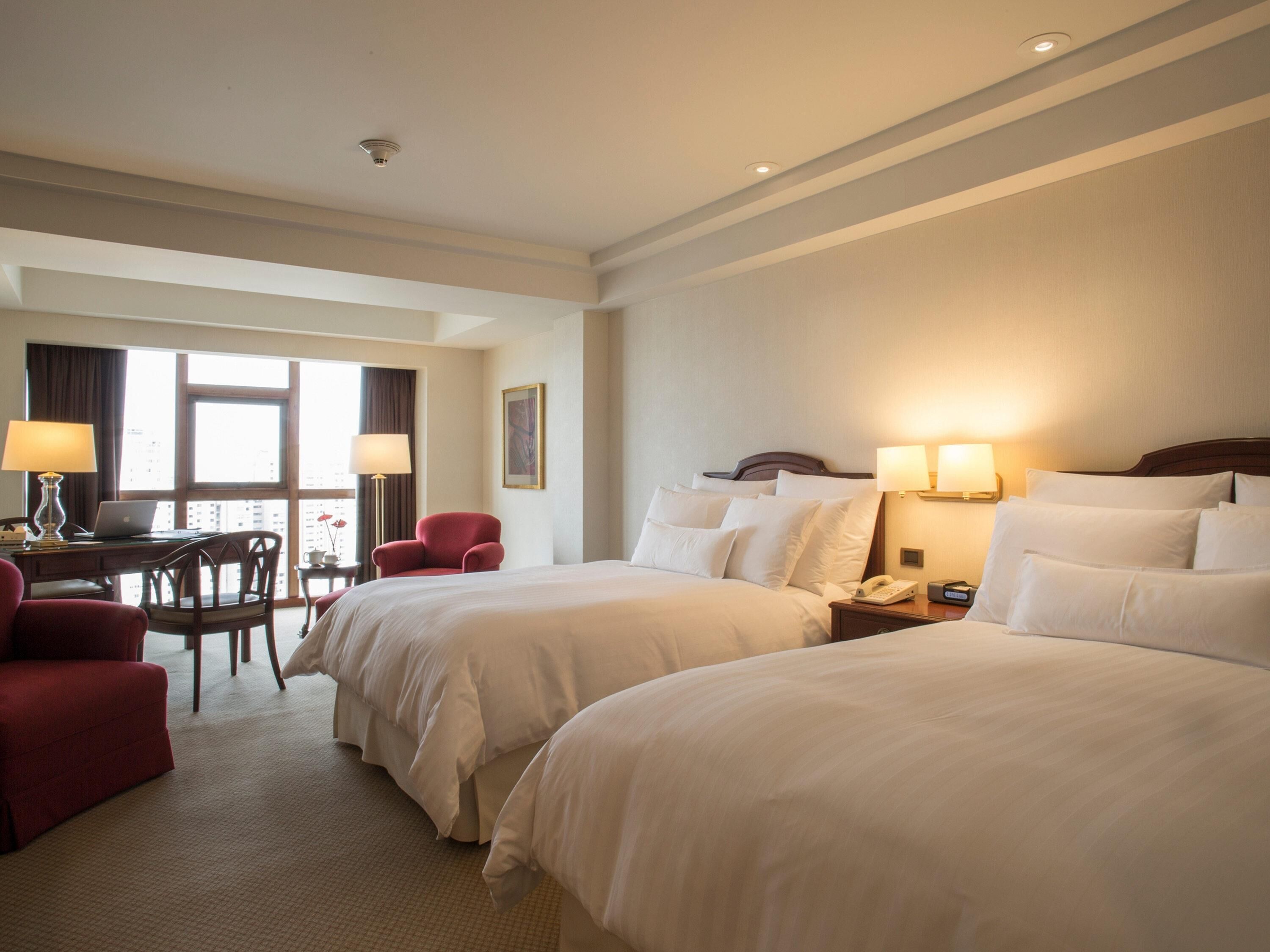 A hotel room with two beds, a desk with a laptop, a chair, and a window with curtains.