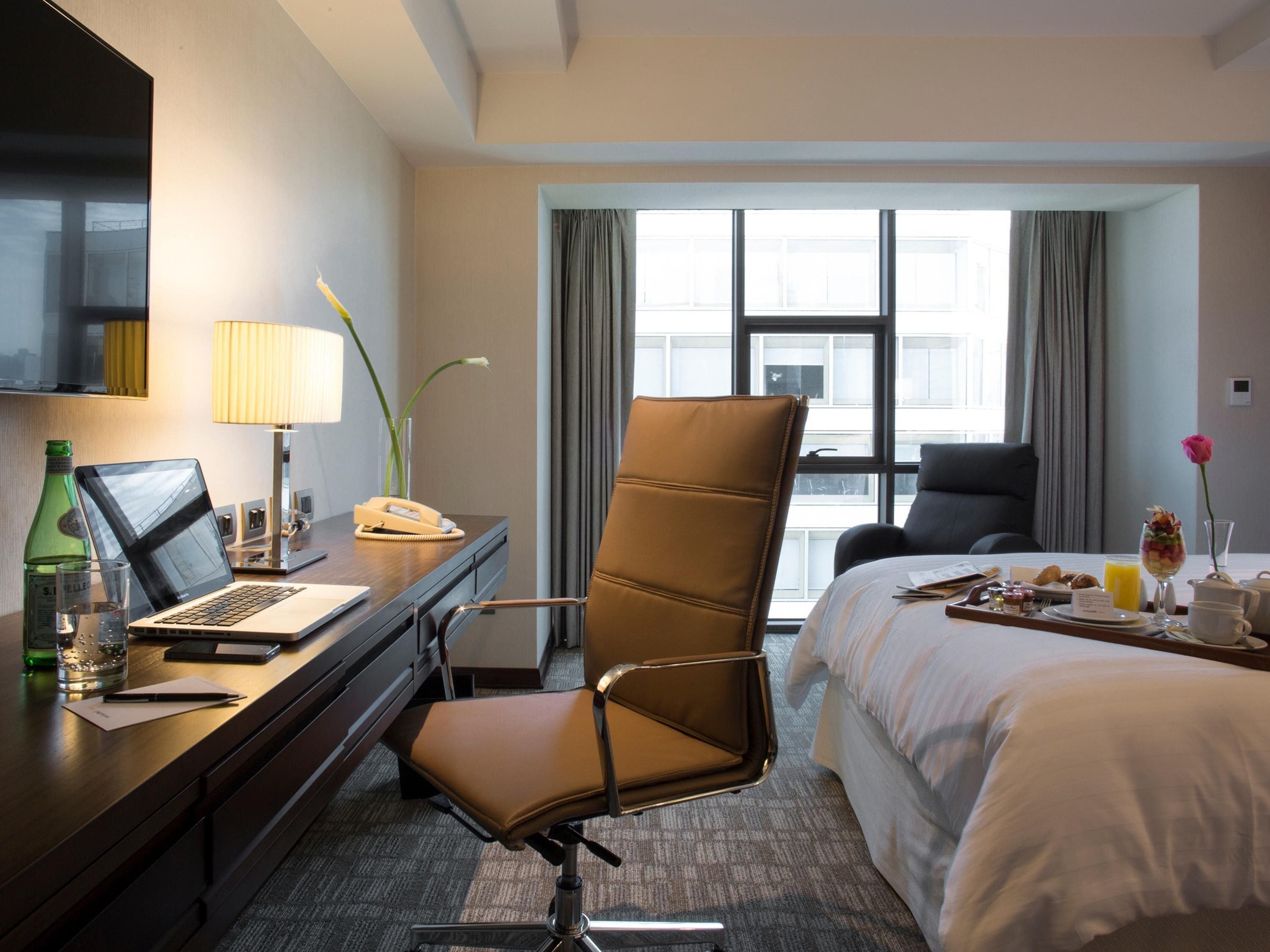 Habitación de hotel con cama, escritorio con computadora portátil, silla, ventana con cortinas y bandeja de desayuno.