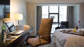 Habitación de hotel con cama, escritorio con computadora portátil, silla, ventana con cortinas y bandeja de desayuno.