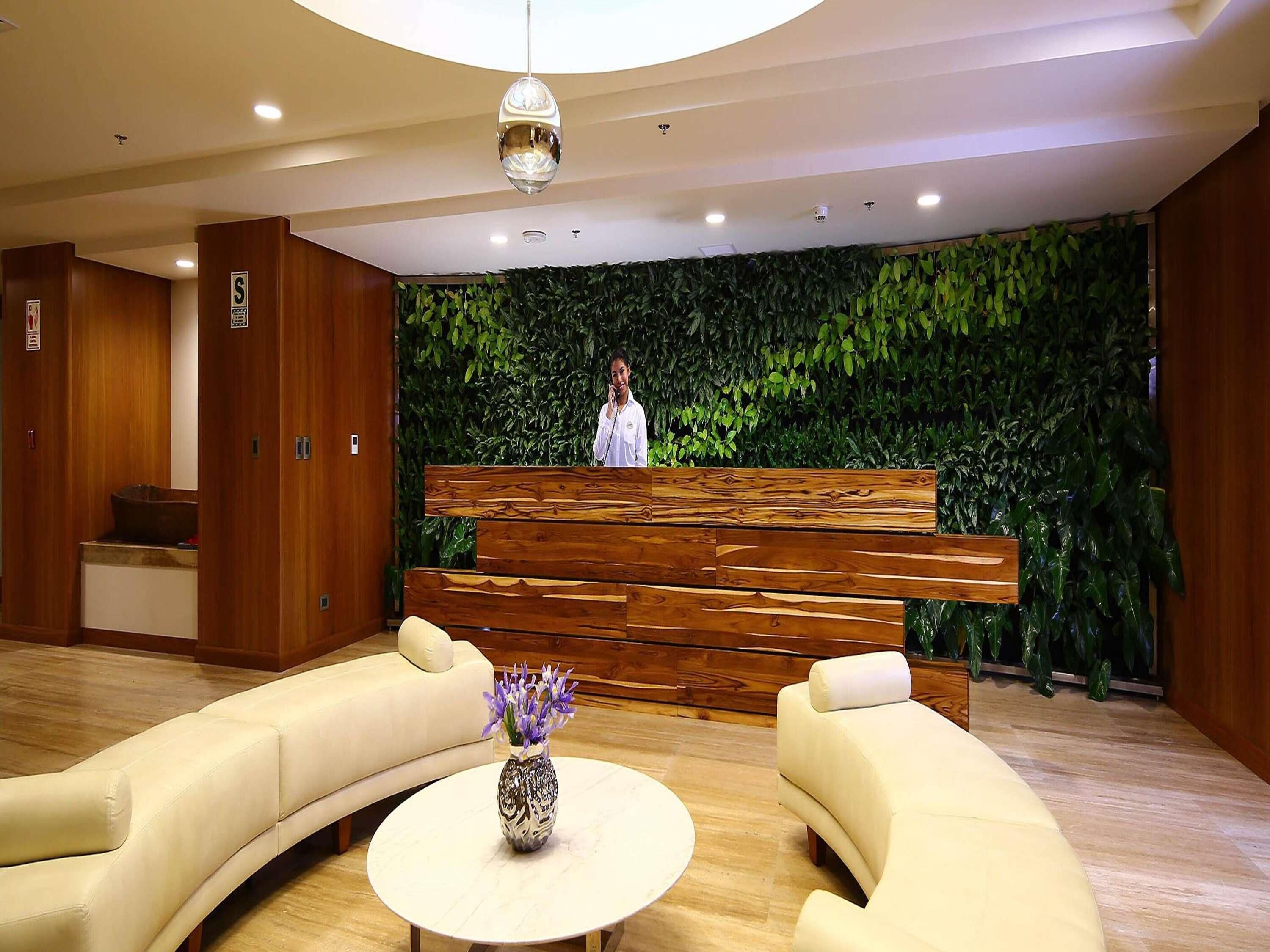 A reception area with a wooden bench, a white sofa, a round table with a vase, and a green wall with plants.