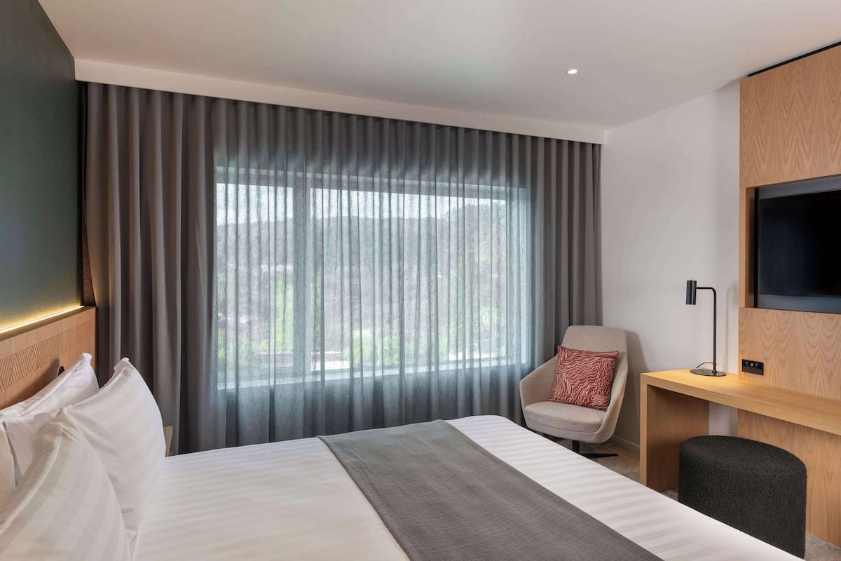 A hotel room with a bed, a chair, a TV mounted on the wall, and a large window with curtains.