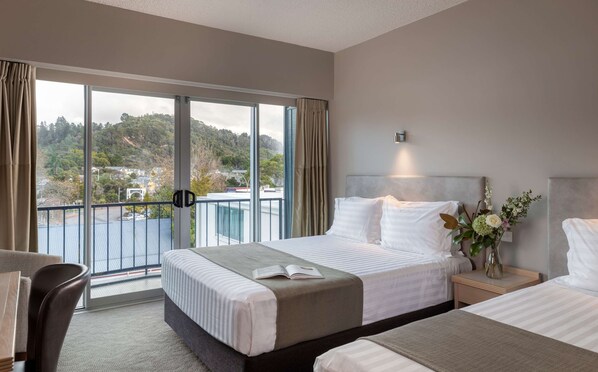 A hotel room with a large bed, a bedside table with a lamp, a view of a balcony, and a window with curtains.