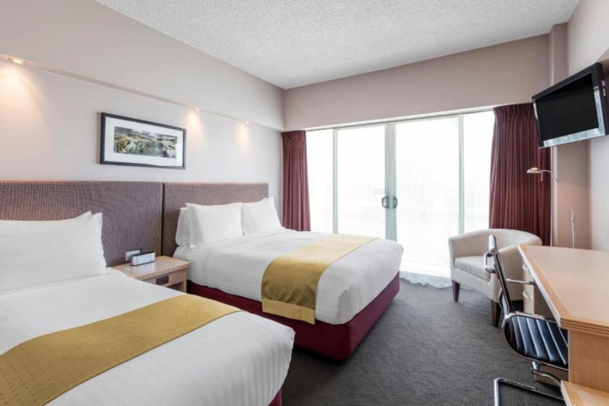 A hotel room with two beds, a desk, a chair, a TV, and a large window.