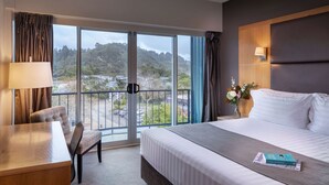 A hotel room with a large bed, a desk, a chair, a nightstand with a lamp, and a view of a town and hills through a balcony. - Rydges Rotorua (Rotorua)