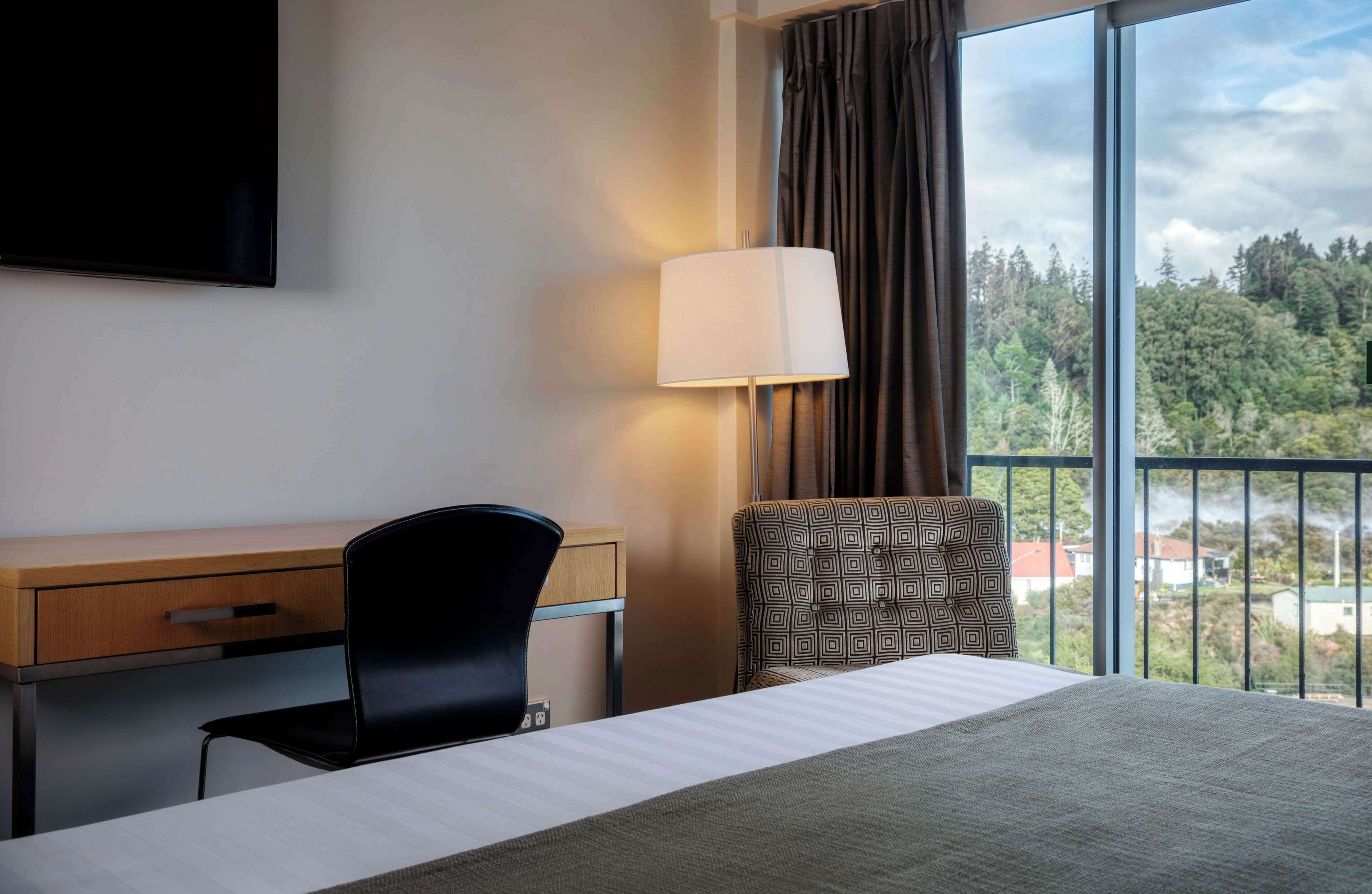 Une chambre d’hôtel équipée d’un lit, d’un bureau, d’une chaise, d’une lampe et d’une grande fenêtre donnant sur des arbres et des immeubles.