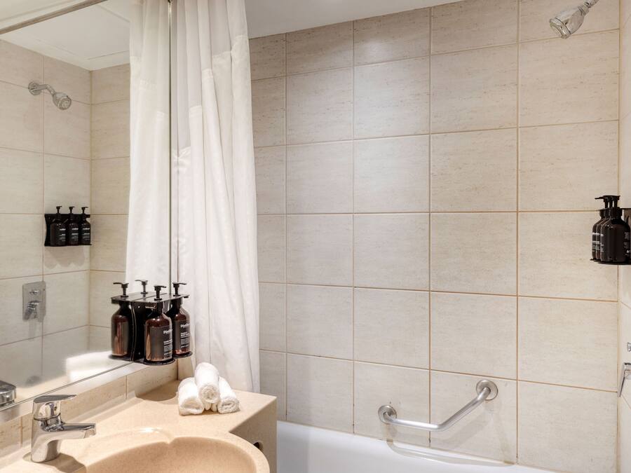 Et moderne badeværelse med en beige flisevæg, et håndvask med spejl, en bruseniche med bruseforhæng og to shampoo flasker på væggen.