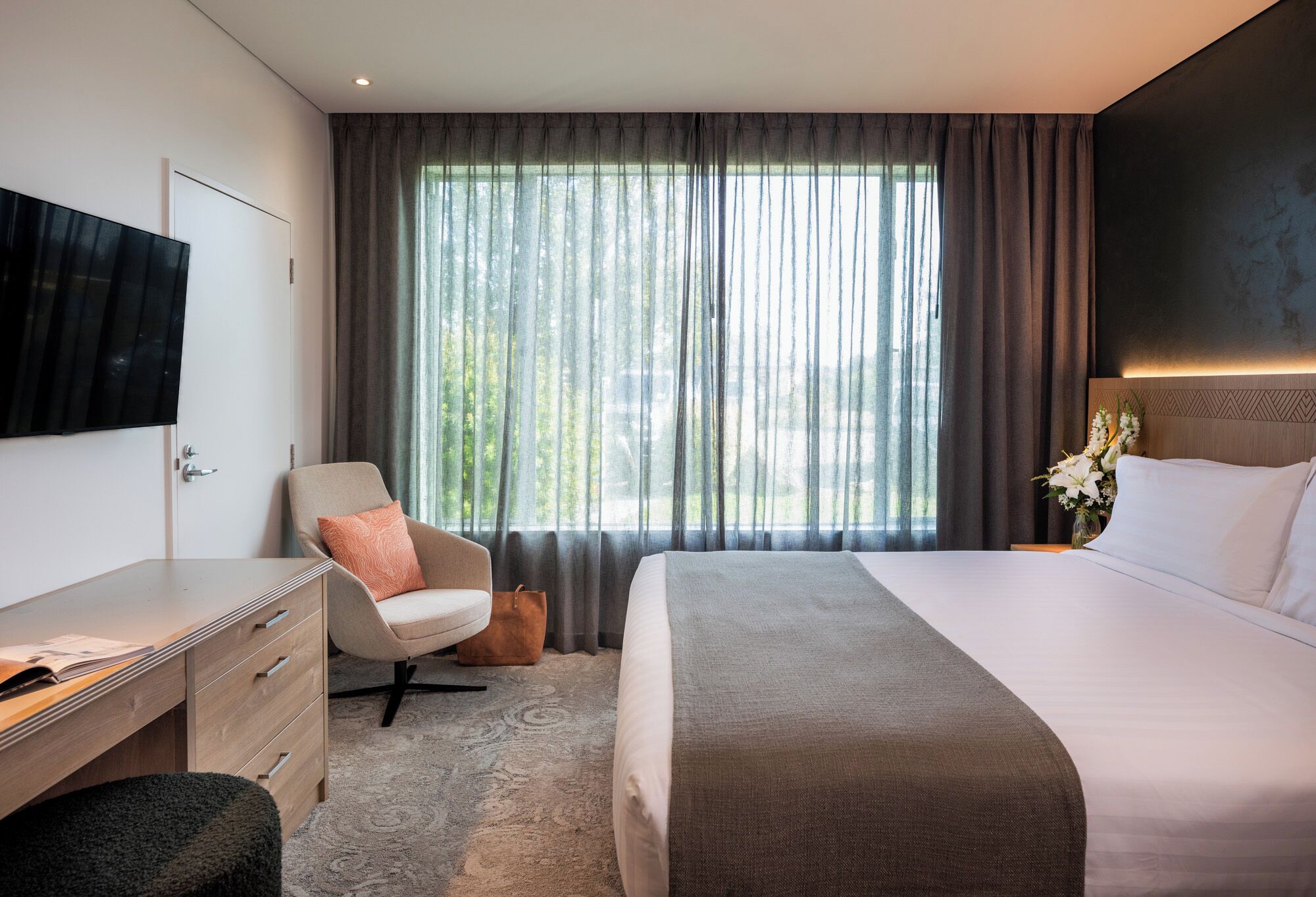 A modern hotel room with a bed, a desk, a chair, and a TV on the wall.
