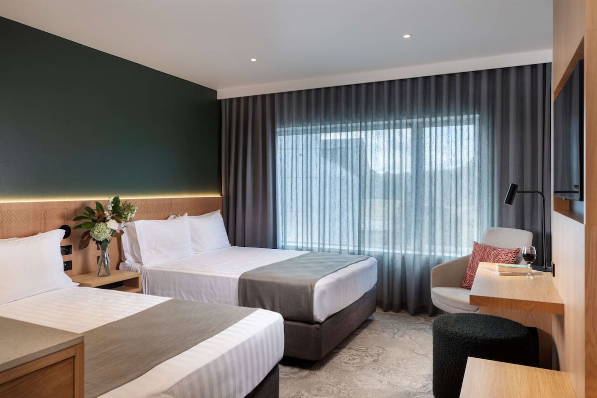 A hotel room with two beds, a desk, a chair, a small table with a wine glass, and a large window with curtains.