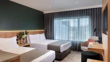 A hotel room with two beds, a desk, a chair, a small table with a wine glass, and a large window with curtains.