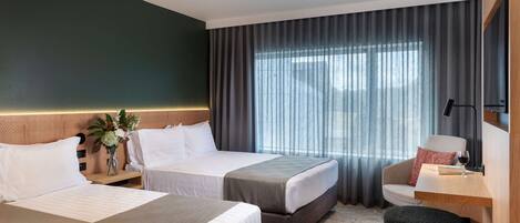 A hotel room with two beds, a desk, a chair, a small table with a wine glass, and a large window with curtains.