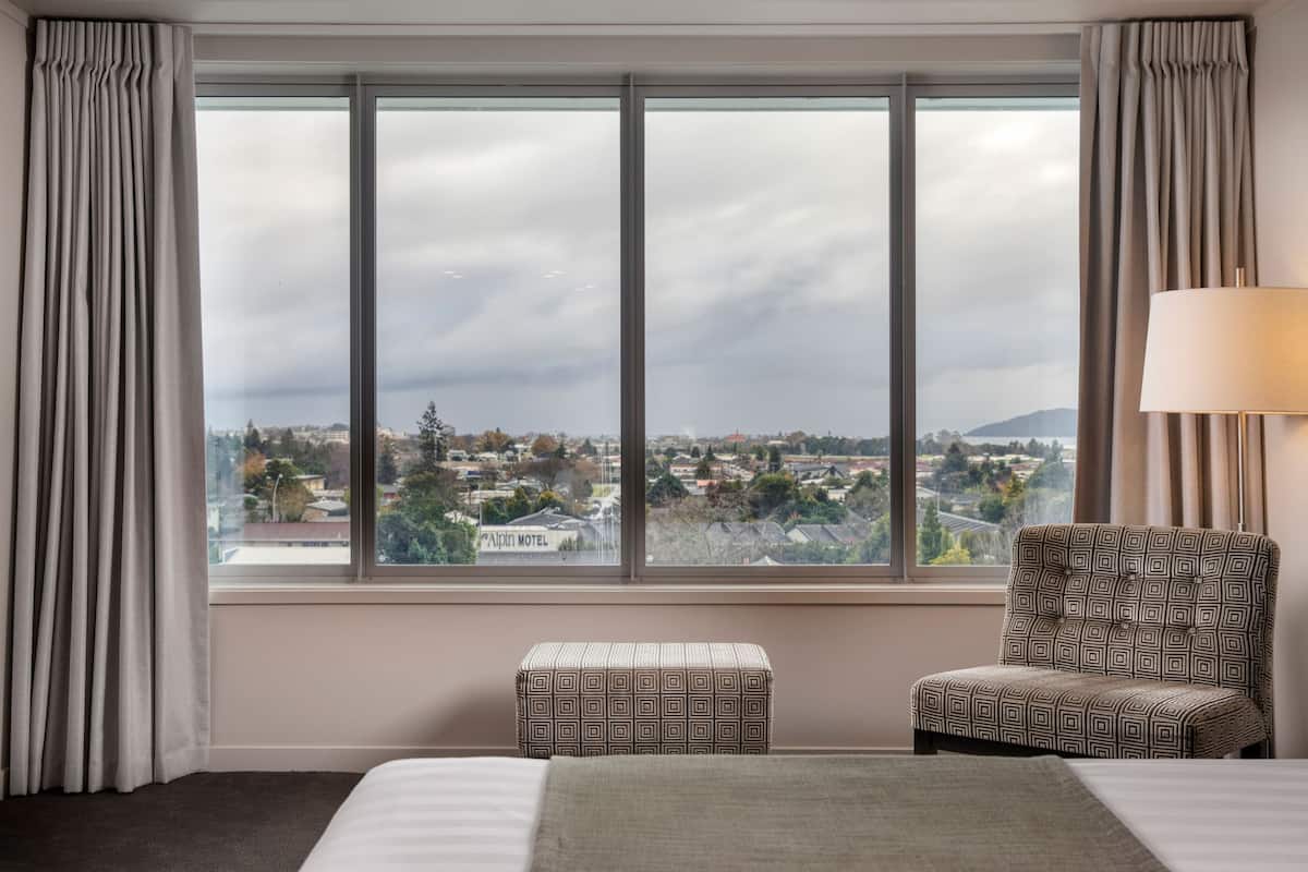 A hotel room with a large window offering a city view, a bed, a patterned armchair, and a small ottoman.