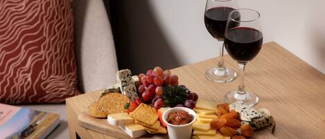 A wooden board with cheese, crackers, grapes, and a bowl of jam on a table. Two glasses of red wine are also visible.