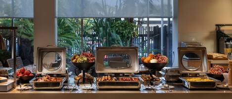 A buffet setup with various food items and a view of a tropical outdoor area through large windows.