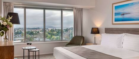 Une chambre d’hôtel avec une grande fenêtre, un lit, une chaise, une petite table avec des verres et une lampe.