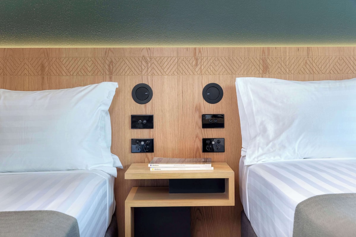 A hotel room with two beds, a small wooden nightstand, and a wall-mounted control panel.