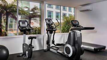 A gym with three cardio machines, a treadmill, and a rowing machine, with large windows offering a view of palm trees and a building.