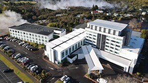 A modern building with a parking lot and a view of a forested area with smoke rising in the background. - Rydges Rotorua (Rotorua)