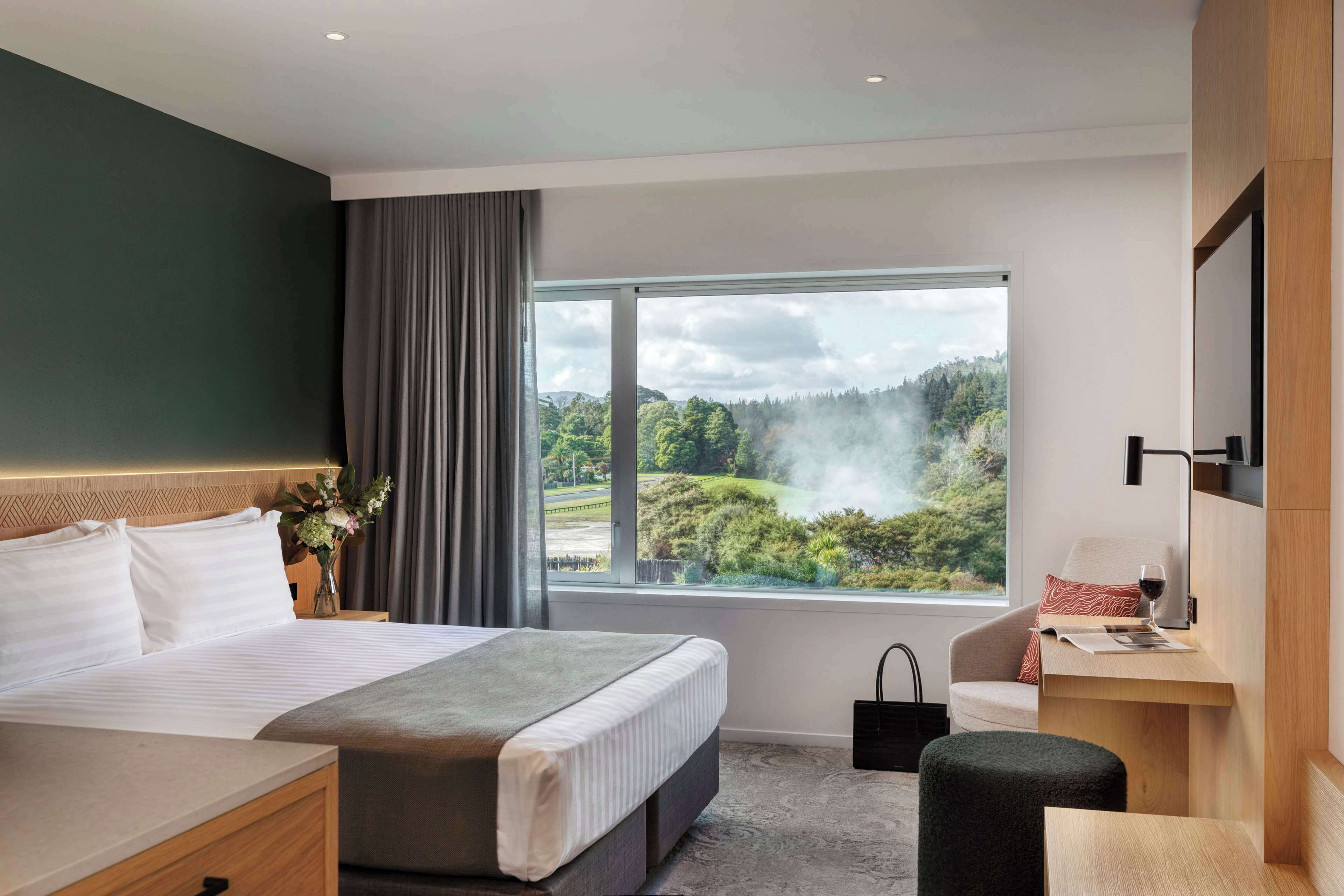 A modern hotel room with a large bed, a desk, and a view of a forested landscape.