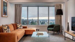 A modern living room with a large window, a sofa, a green armchair, a coffee table with books and glasses, and a TV on a stand. - Rydges Rotorua (Rotorua)