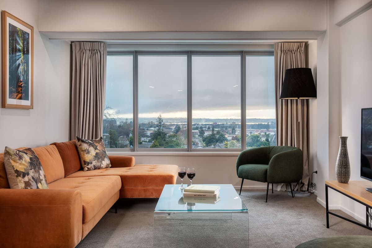 A modern living room with a large window, a sofa, a green armchair, a coffee table with books and glasses, and a TV on a stand.