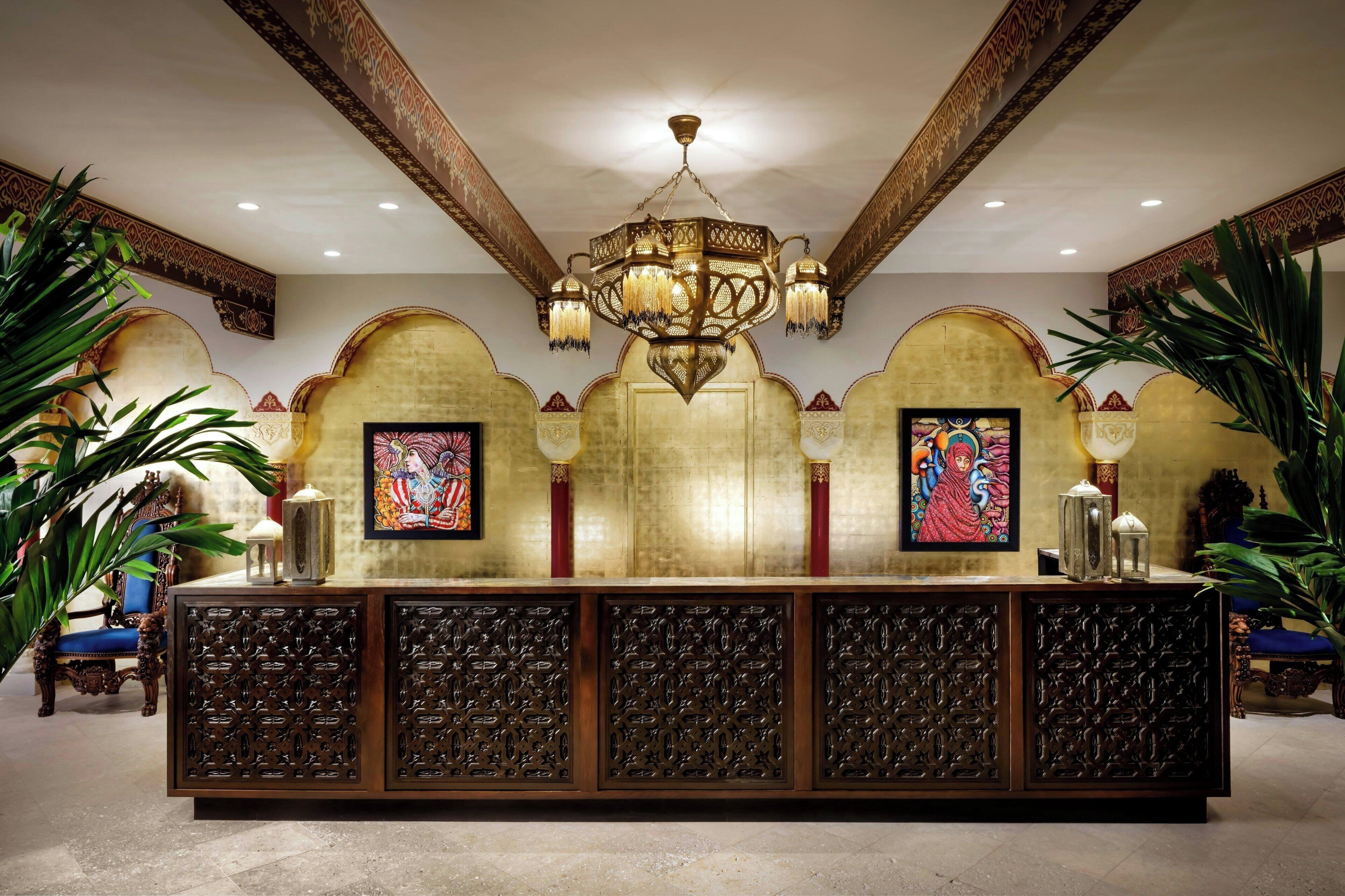 A reception area with a large, ornate chandelier, a reception desk with intricate carvings, and framed artwork on the walls.
