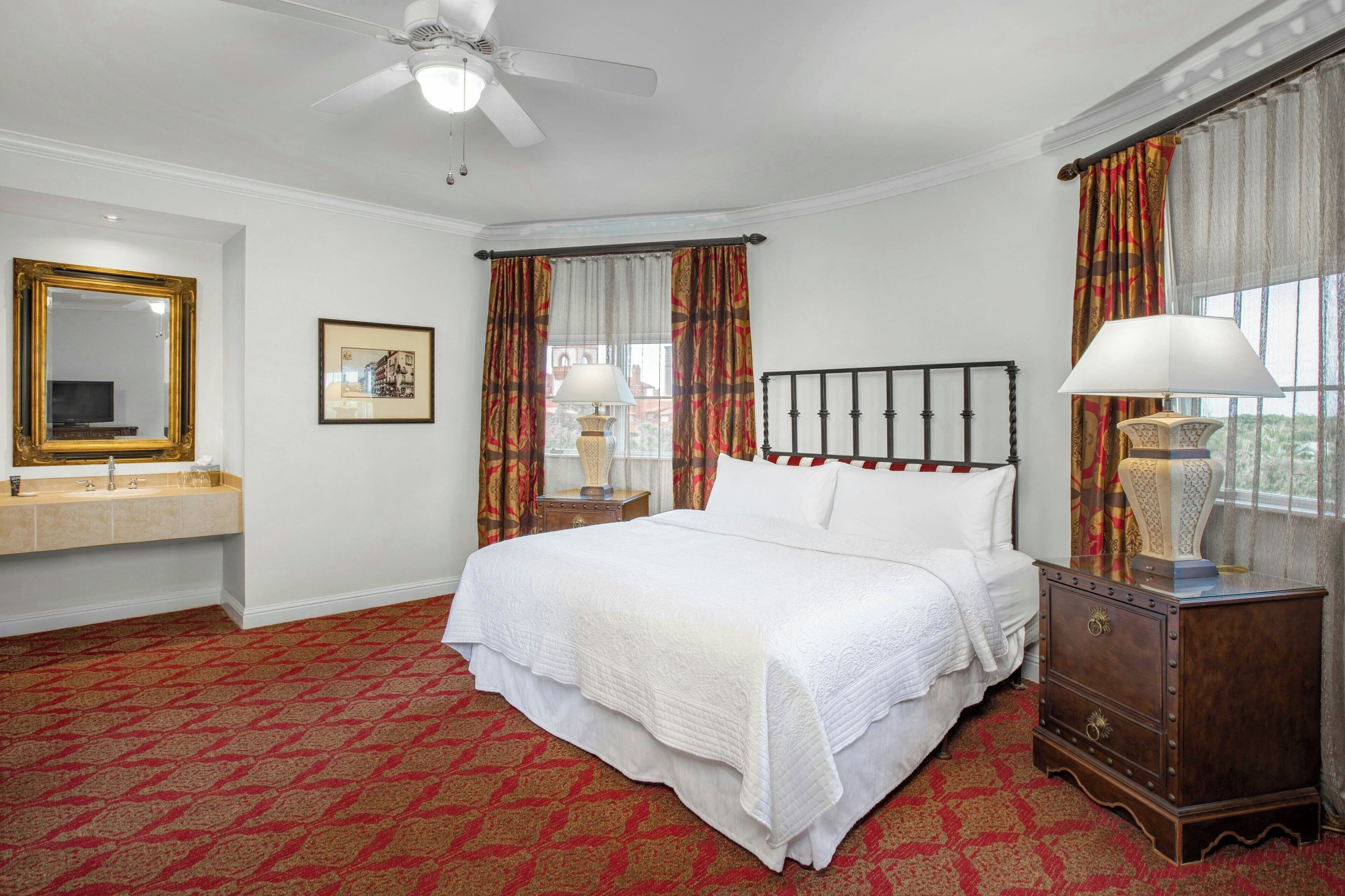 Une chambre à coucher comprenant un lit, une table de chevet, un miroir, une commode et un ventilateur de plafond.