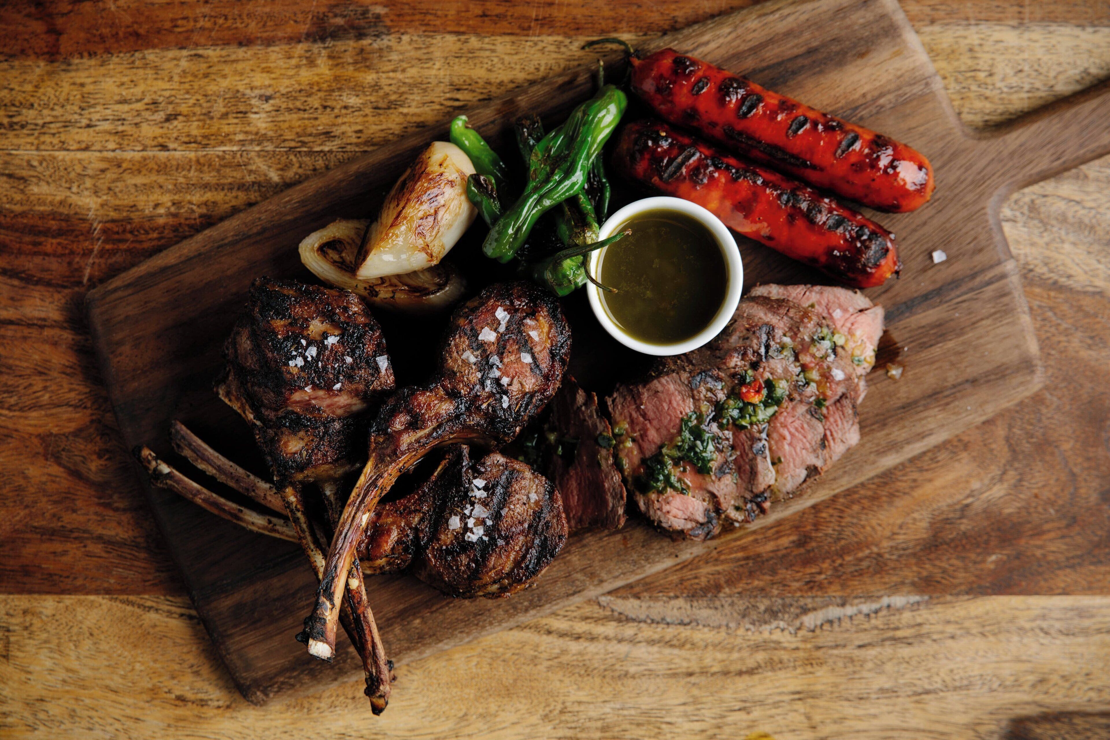 A wooden cutting board with various meats, including grilled sausages, roasted potatoes, and a piece of steak, accompanied by a small bowl of sauce.