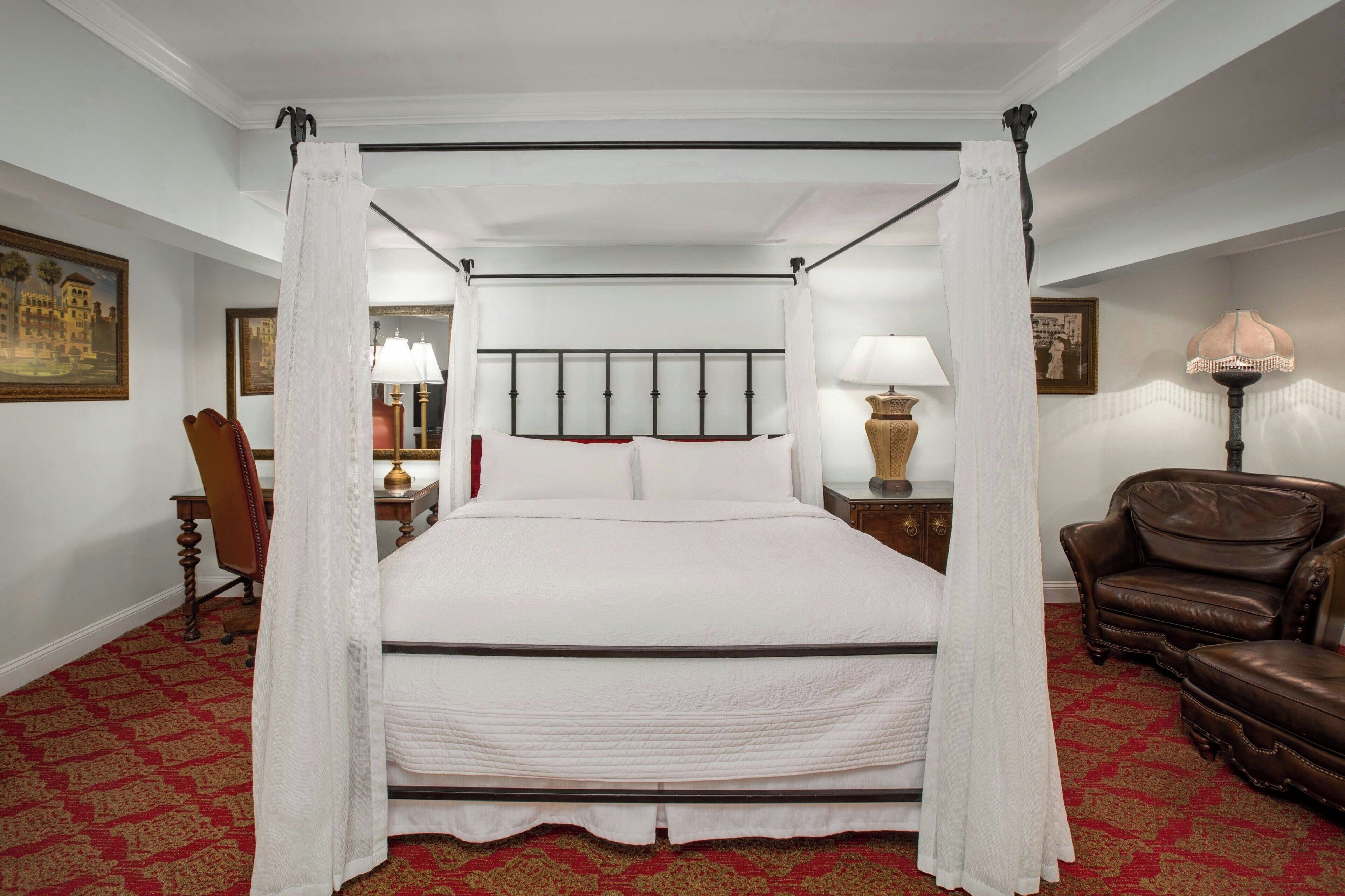 A four-poster canopy bed with white bedding, a red patterned carpet, and a brown leather armchair.