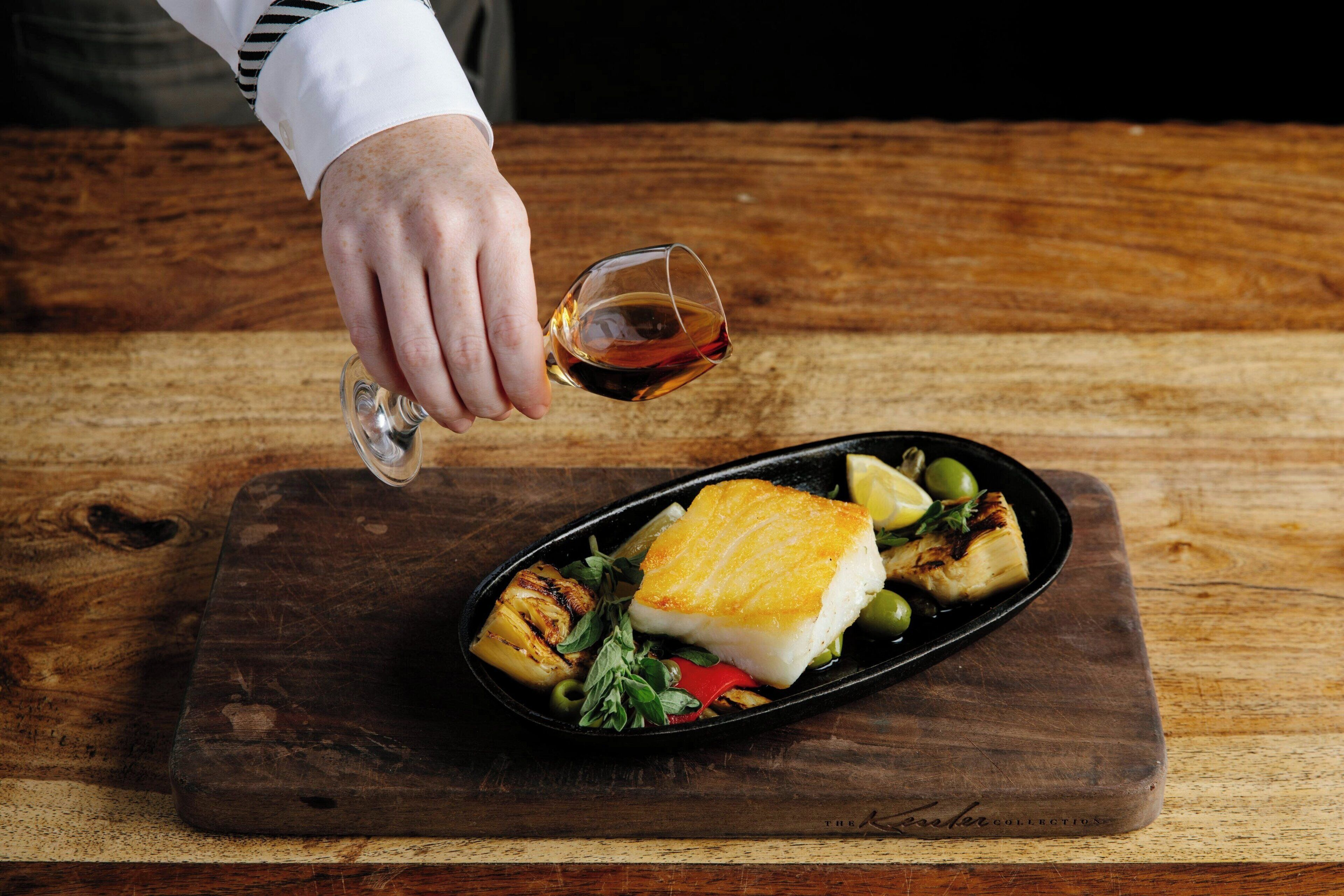A chef pouring wine onto a dish with fish, vegetables, and a lemon wedge on a wooden board.