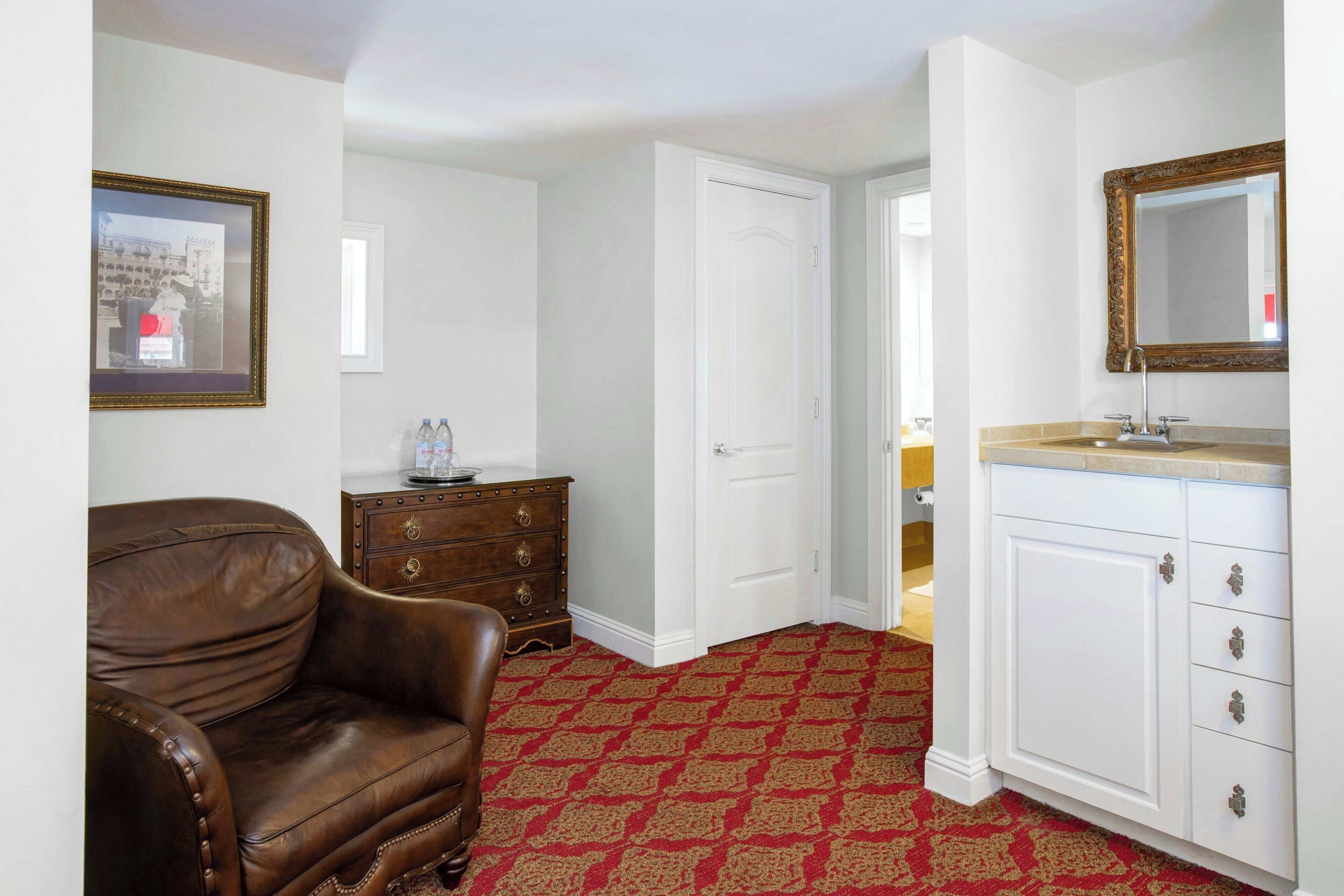 A room with a brown leather sofa, a white door, a white cabinet with a mirror, and a patterned carpet.
