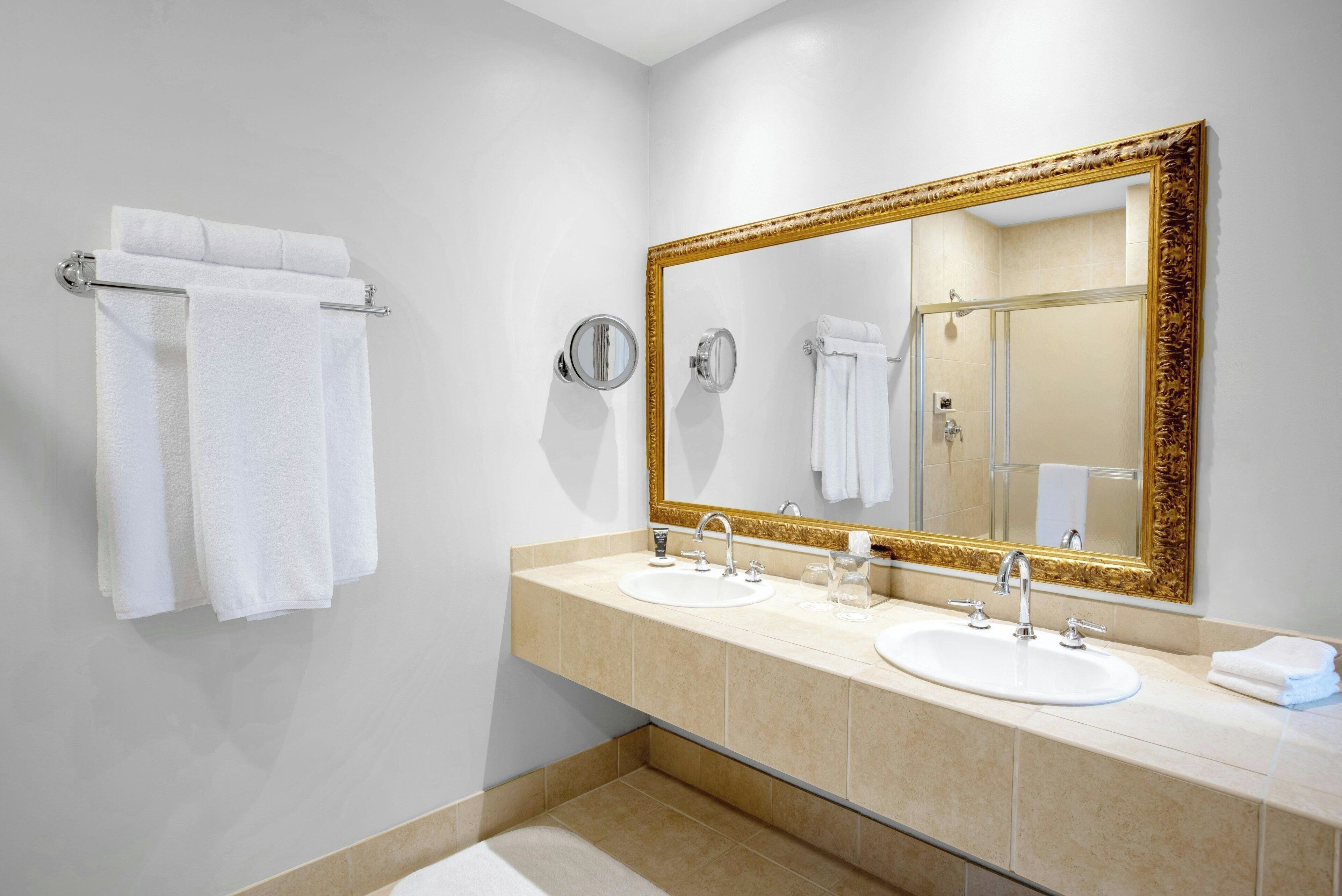 Une salle de bain avec deux lavabos, un grand miroir avec un cadre doré et un porte-serviettes avec des serviettes blanches.