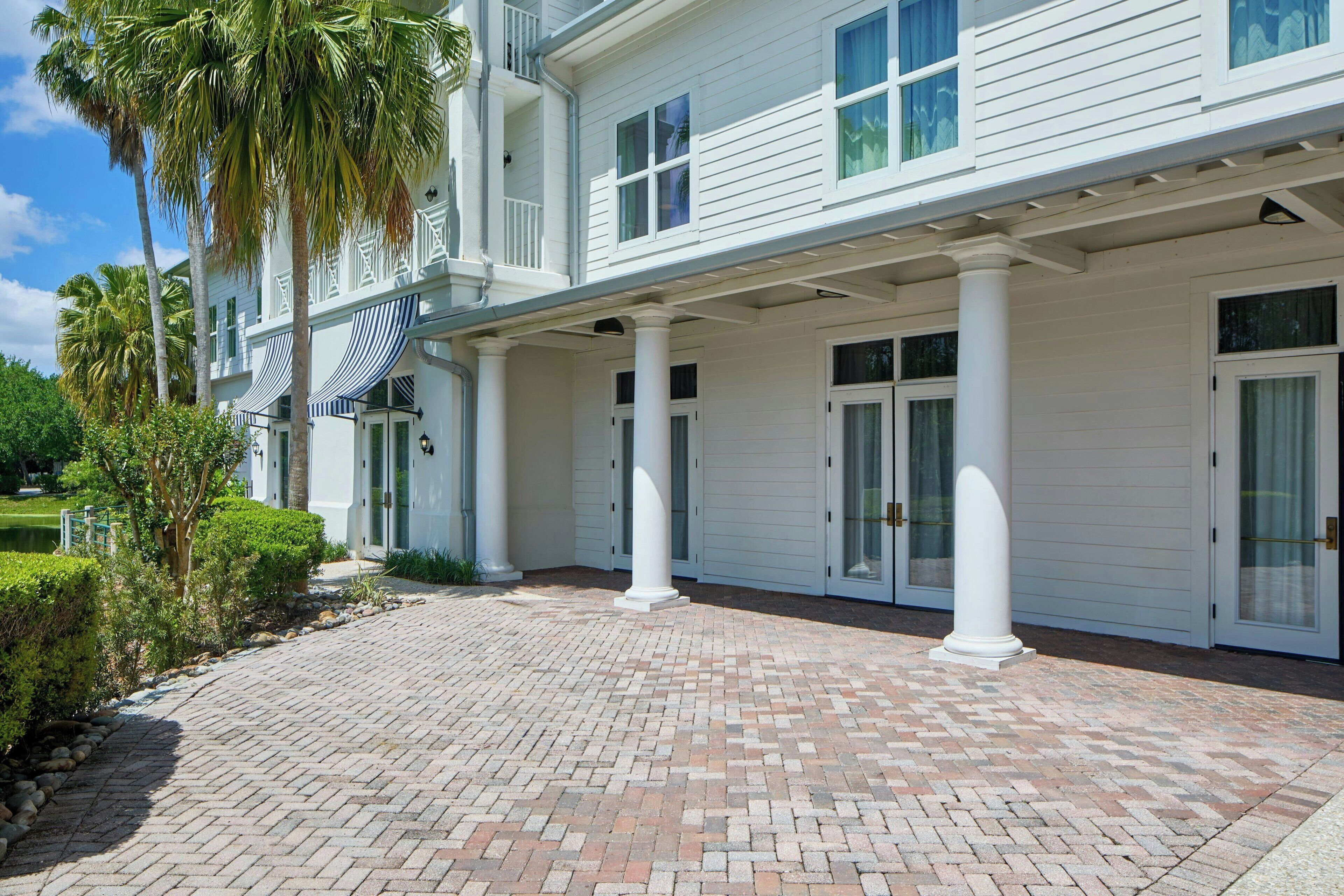 A two-story building with a covered porch supported by white columns, a brick pathway, and palm trees.