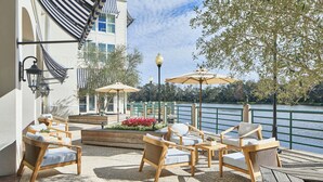 A terrace with wicker furniture, a view of a lake, and a building with a striped awning. - The Inn at Celebration, Autograph Collection  (Celebration)