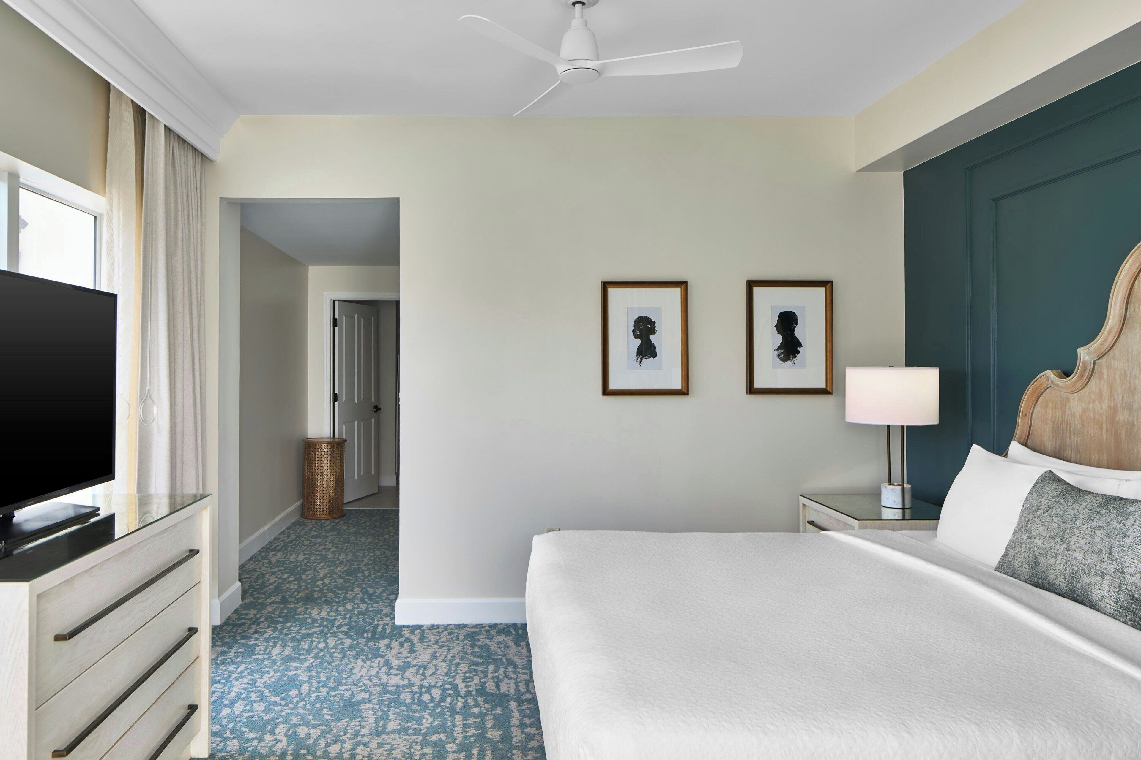 Une chambre d’hôtel équipée d’un lit, d’une télévision, d’un ventilateur de plafond et de deux tableaux encadrés accrochés au mur.