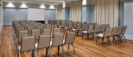 Un salón de conferencias con filas de sillas, una pantalla para proyector y un ventanal con cortinas.