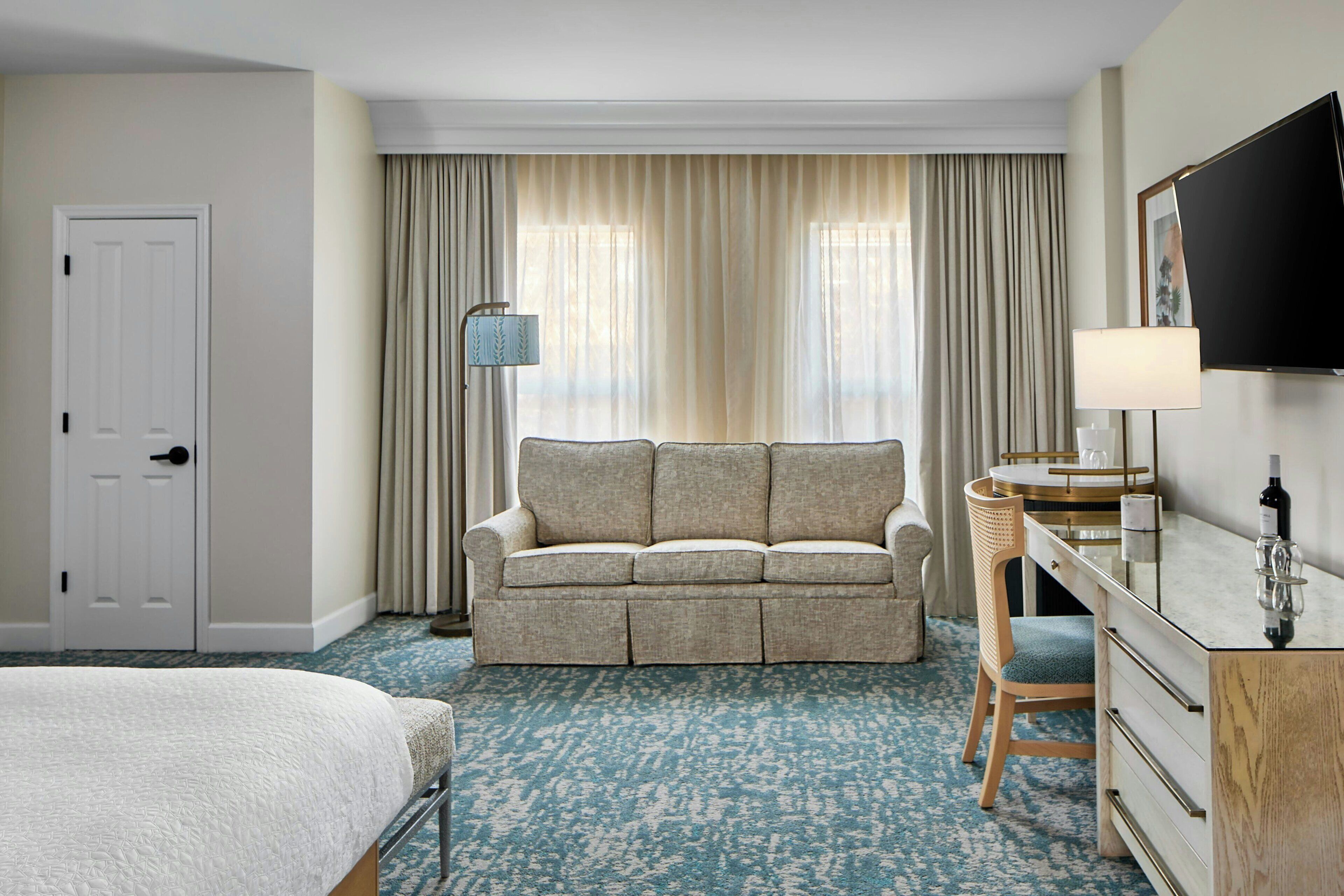 Habitación de hotel con cama, sofá, escritorio con silla, televisor y ventana con cortinas.