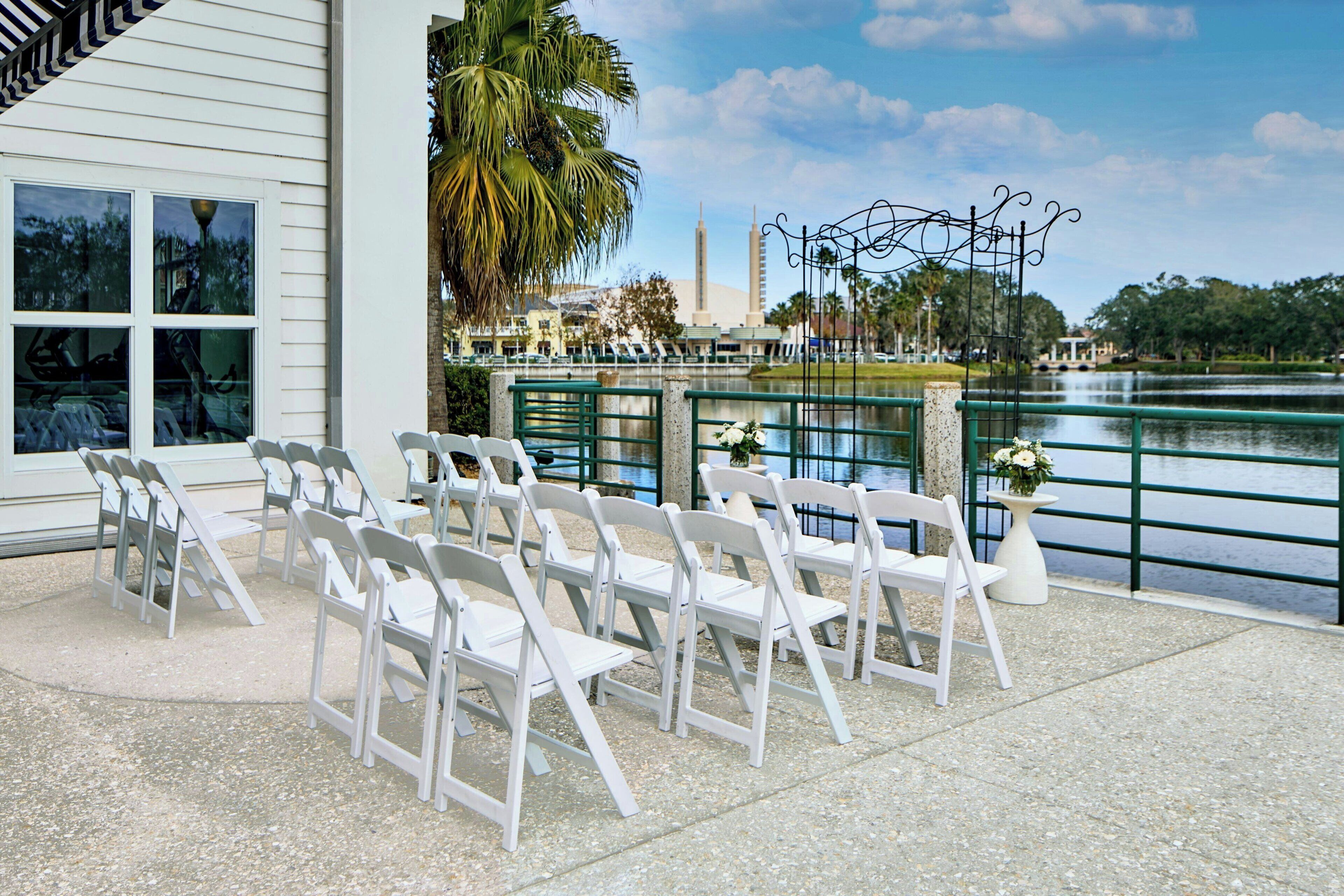 Outdoor seating with white chairs arranged on a concrete patio overlooking a lake, with a building and palm trees in the background.