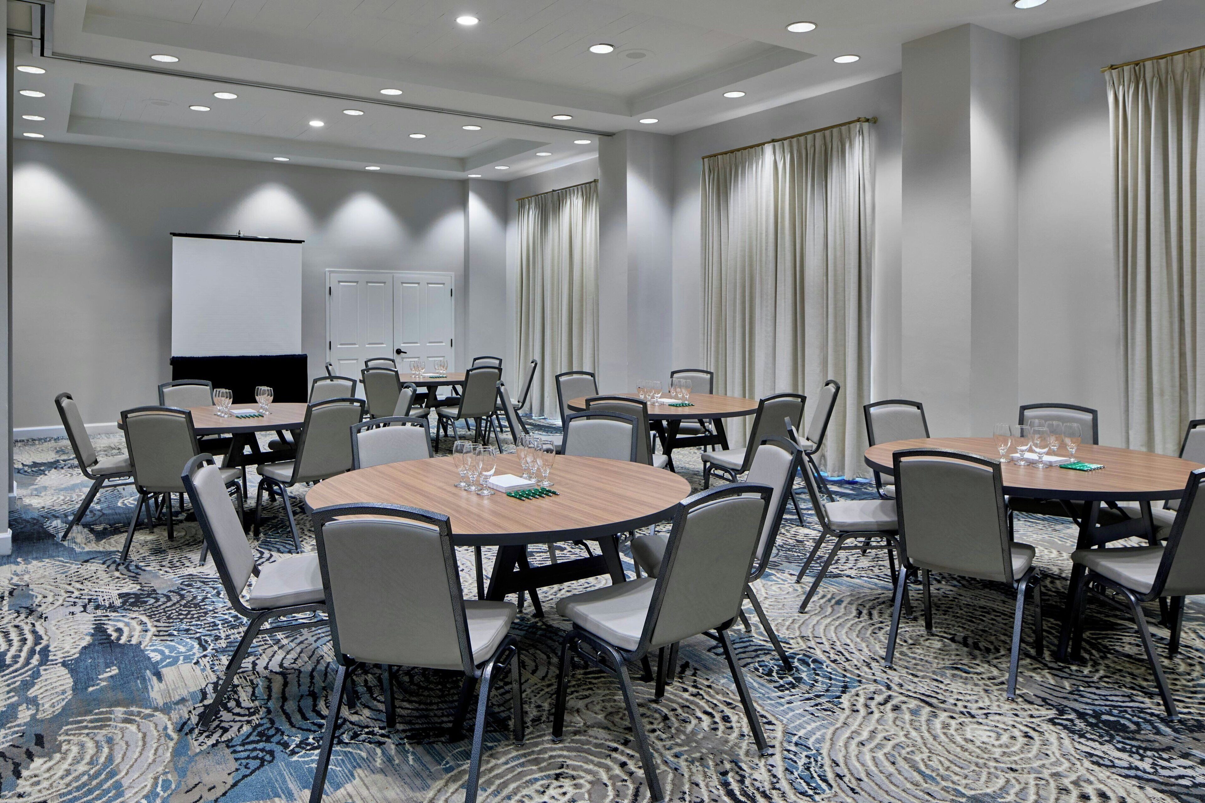 A conference room with multiple round tables, chairs, a whiteboard, and a projector screen.