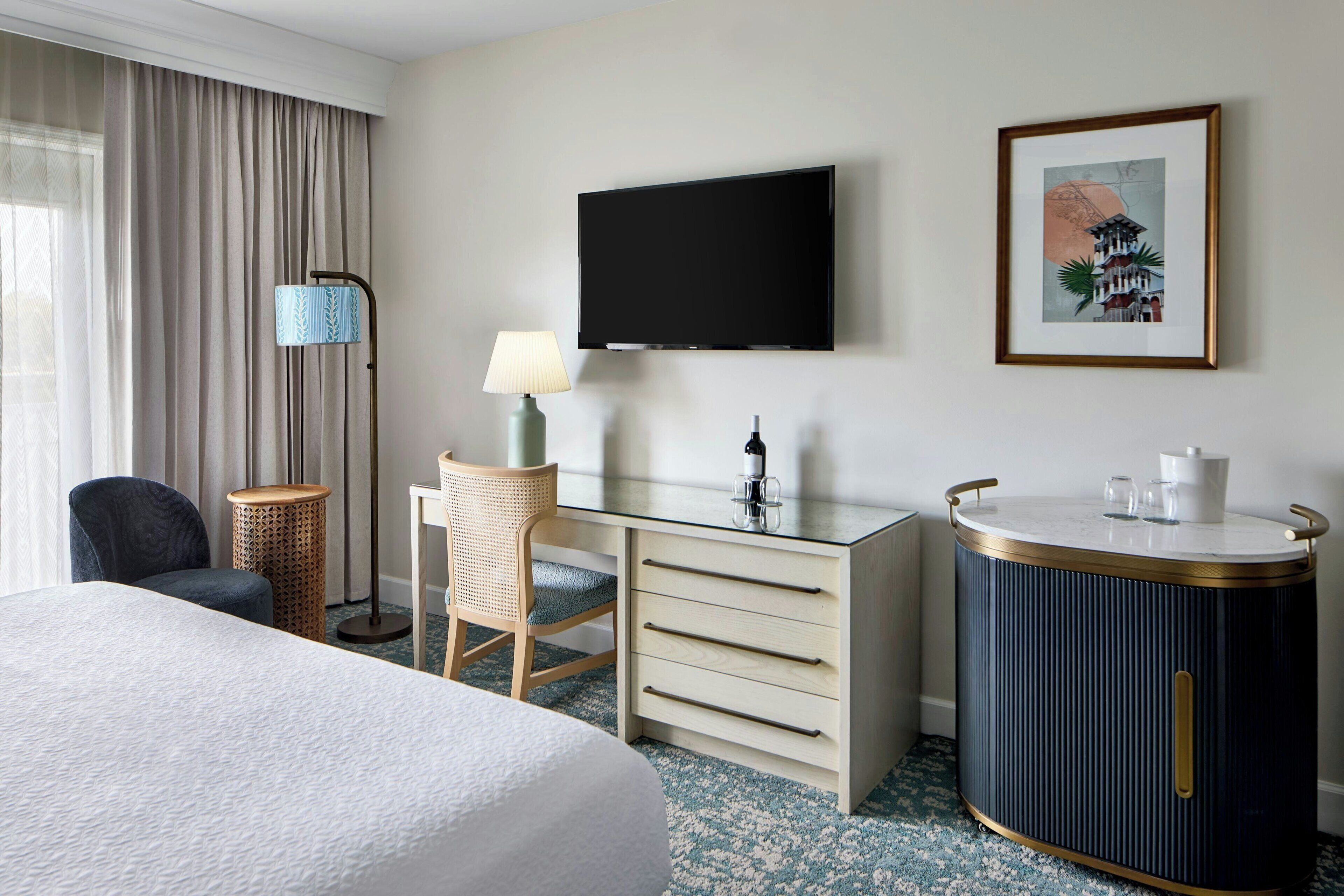 A hotel room with a bed, a desk with drawers, a television, a lamp, a chair, a round cabinet, and a framed picture on the wall.