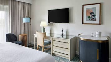 Habitación de hotel con cama, escritorio con cajones, televisor, lámpara, silla, armario redondo y un cuadro en la pared.