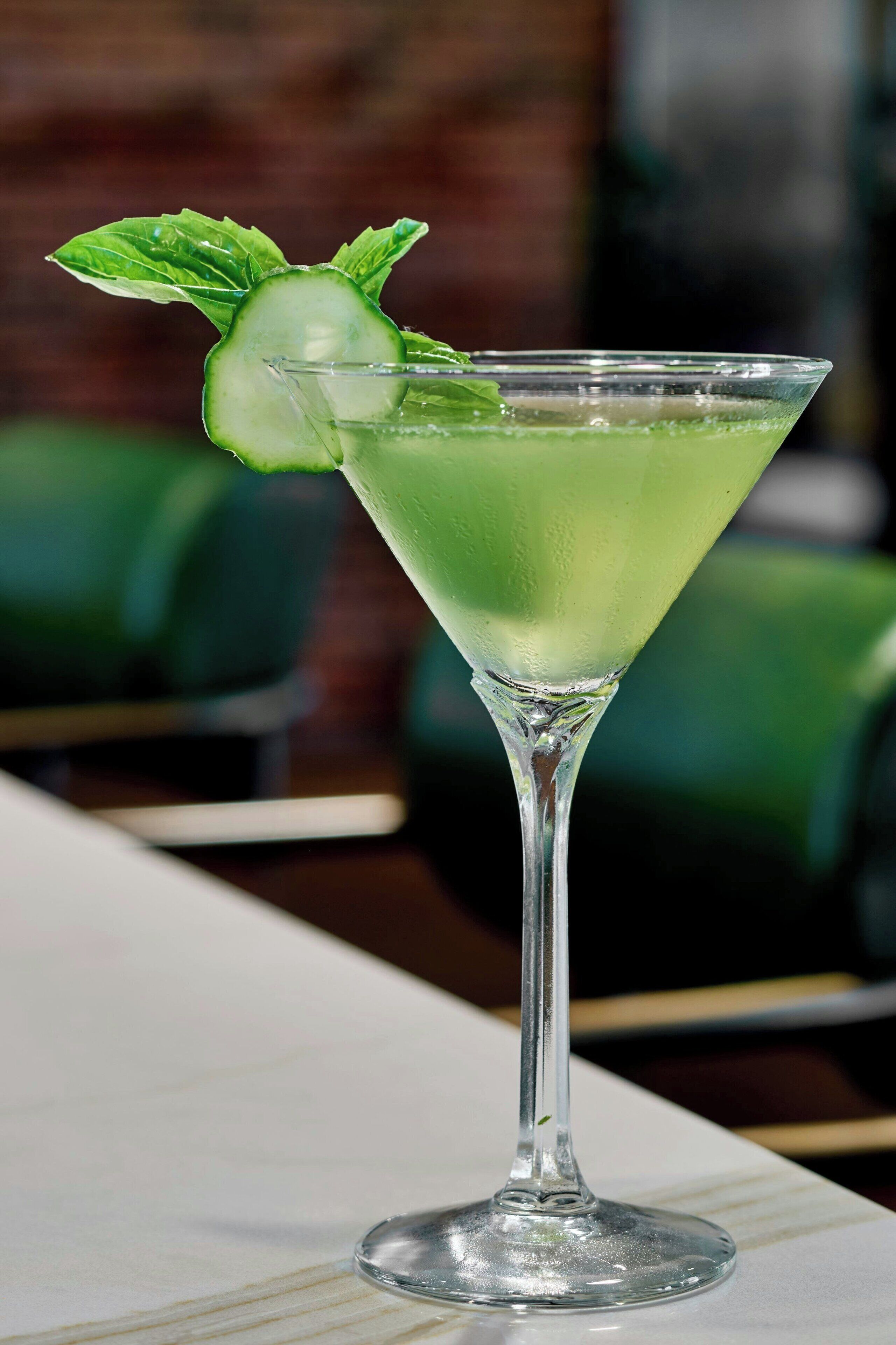 A martini glass with a green drink, garnished with cucumber slices and mint leaves, on a white table.